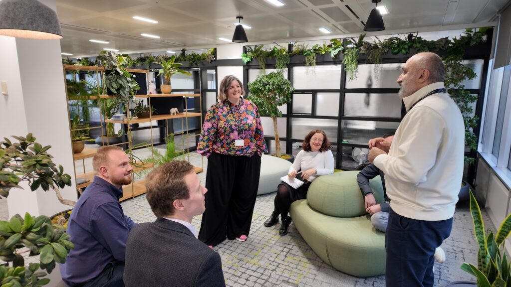 A group of Aberdeen Group employees engaged in a casual meeting in a modern, plant-filled office space.