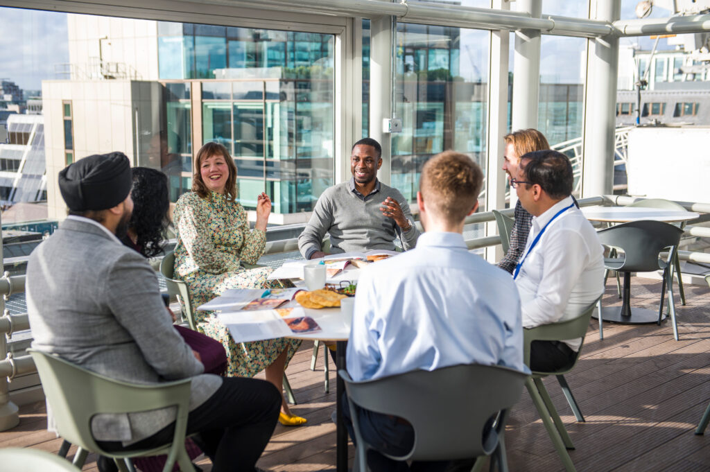 A diverse group of AXA UK colleagues sitting around a circular table in a bright, glass-walled rooftop office space. They are engaged in a lively discussion with documents and refreshments on the table, while a city skyline is visible in the background