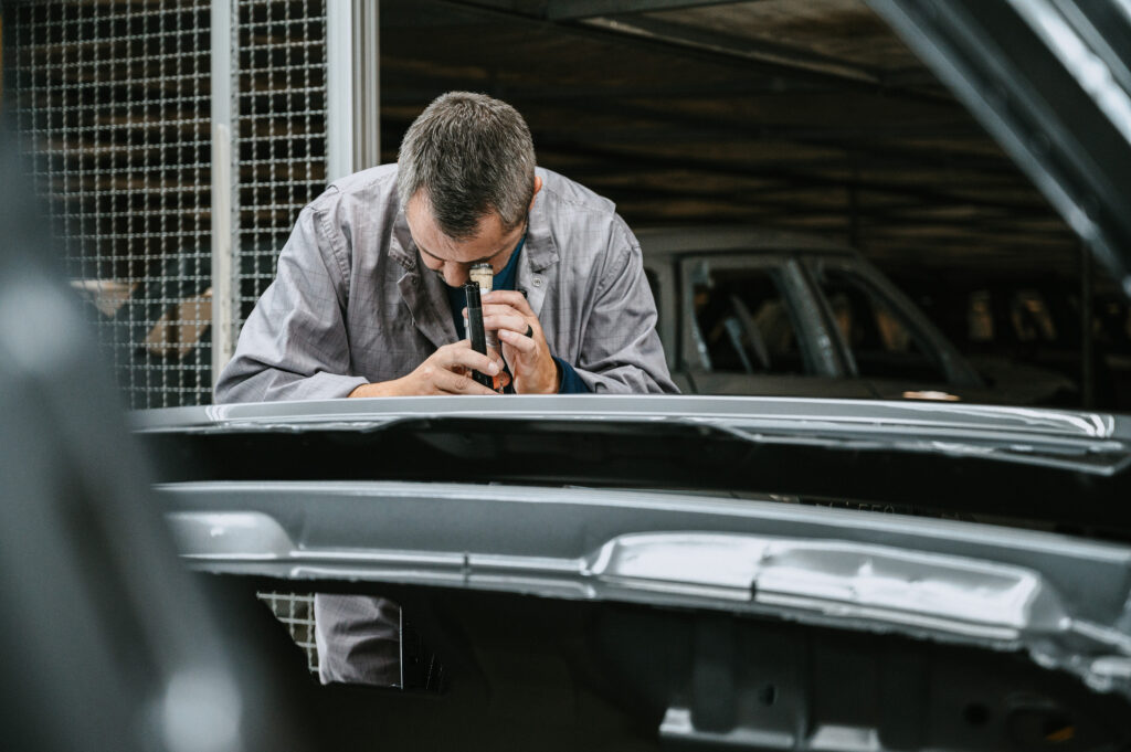 An JLR automotive technician in a grey work coat looking closely through a specialised magnifying loupe tool to inspect the metallic grey paint finish of a car chassis.