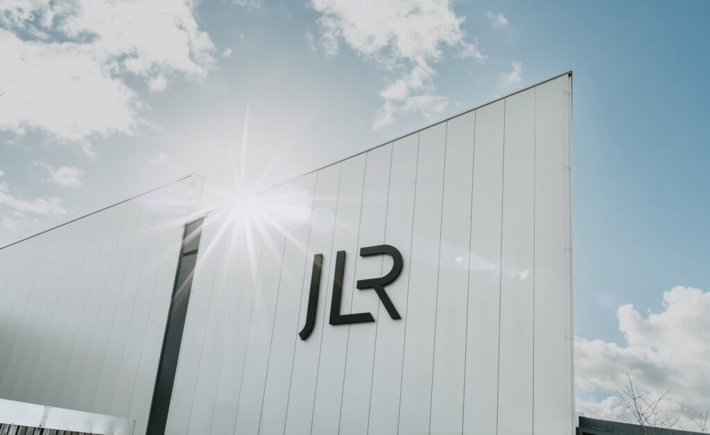 The minimalist black JLR corporate logo mounted on a large, modern white paneled exterior building wall under a bright blue sky with sun flare.