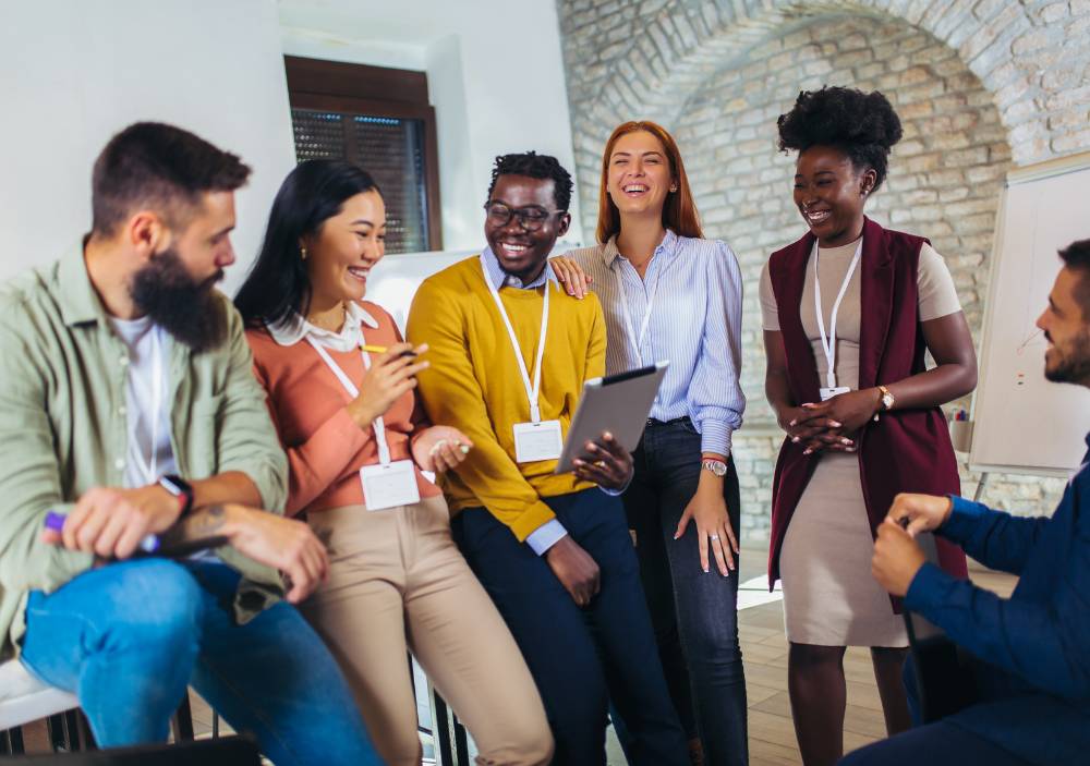 A diverse group of six young professionals smiling and collaborating during a workshop or meeting, with one holding a tablet.